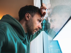 Bild mti einem mittelalten Mann, der traurig schaut und der mit Hand und Kopf an einem Fenster lehnt
