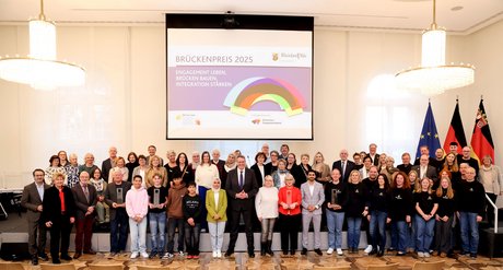 Gruppenfoto aller Ausgezeichnetetn mit Ministerpräsident Alexenander Schweitzer vor dem Logo mit Brückenpreis 2025