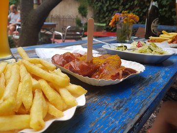 Currwurst, Pommes Bierflasche auf einer blauen Festgarnitur im Hintergurnd Menschen