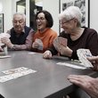 Am Tisch sitzend ein älterer Mann und zwei lachende Frauen halten Spielkarten in der Hand, ein Händepaar im Anschnitt hält ein Kartenblatt in der Hand