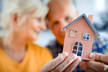 Bild mit älterem Mann und Frau, die ein Modellhaus in ihren Händen halten.