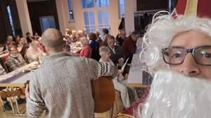 Selfie-Bild eines Mannes der als Nikolaus mit weißem Bart und roter Mütze verkleidet ist, im Hintergrund ein Mann der Gitarre spielt und ältere Menschen an gedeckten Tischen.