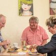 Zwei ältere Männer und eine Frau sitzen an einem Tisch mit Café und Kuchen und unterhalten sich, im Hintergrund einige Bilder an der Wand