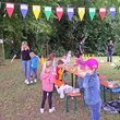 Bierbank auf Wiese unter Bäumen an denen bunte Wimpel hängen. Kinder und Erwachsene sind bei diversen Spielen auf der Wiese