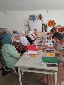 Bild auf dem Frauen mit und ohne Kopftüchern zu sehen sind. Sie sitzen an einem langen Tisch mit Kaffeetassen, Heften und Papieren und unterhalten sich.