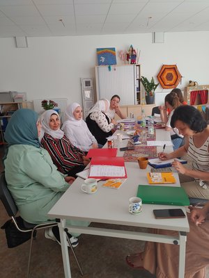 Bild auf dem Frauen mit und ohne Kopftüchern zu sehen sind. Sie sitzen an einem langen Tisch mit Kaffeetassen, Heften und Papieren und unterhalten sich.