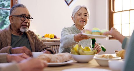 Senioren sitzen am gedeckten Frühstückstisch, eine Frau reicht einer anderen einen Teller mit Cup-Cakes