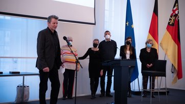 Mann am Mikrofon bei der Dankesrede, im Hintergrund vor fünf Personen mit Masken