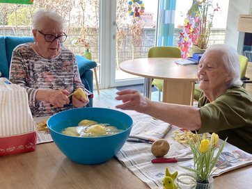 2 alte Damen sind über Eck am Tisch und schälen Kartoffeln, die geschälten Kartoffeln schwimmen in einer blauen Schüssel mit Wasser auf der Tischmitte.
