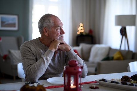 alter Mann, der alleine mit aufgestütztem Kopf und traurig an einem Tisch sitzt, im Hintergrund Weihnachtsdekoration