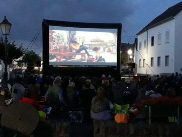 In der Dämmerung auf einem Außenplatz im Ort eine große Leinwand auf der ein Film läuft, viele Zuschauer sitzen davor