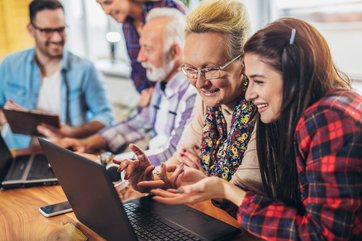 zwei junge Frauen und ein junger Mann erklären einem Senior und einer Seniorin an einem Tisch mit Laptops digitale Anwendungen. 